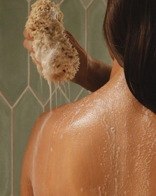 “Person washing their back with a natural bath sponge, creating lather and water drips on skin against a tiled shower background.”