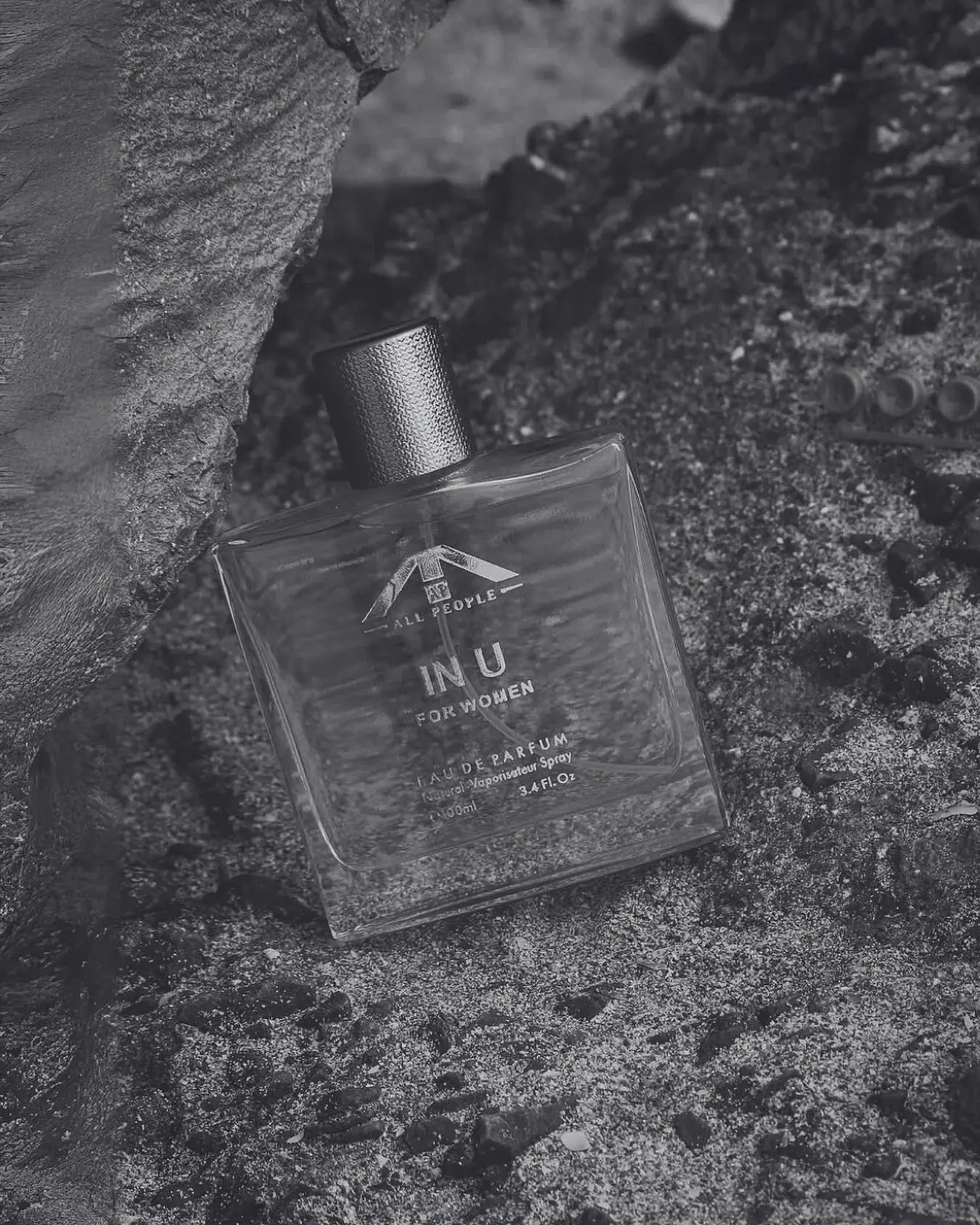 “All People ‘INU’ perfume for women, placed against textured rocks and sand in a monochrome setting, showing a clear glass bottle with a metallic cap.”
