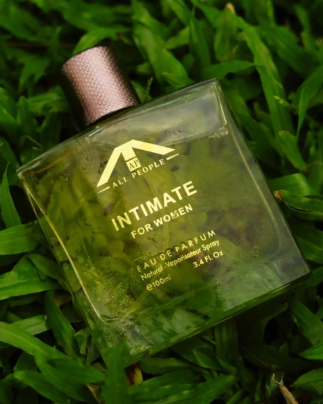 “All People ‘Intimate’ perfume for women, placed on fresh green grass, showing a clear glass bottle with a textured cap in a natural outdoor setting.”