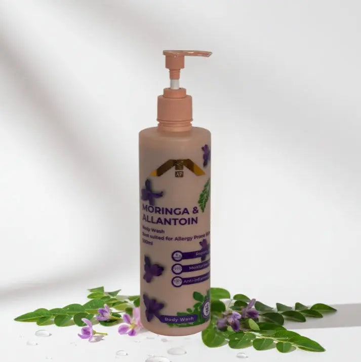“All People Moringa & Allantoin Body Wash bottle with pump, standing among fresh moringa leaves and purple blossoms on a soft white backdrop.”