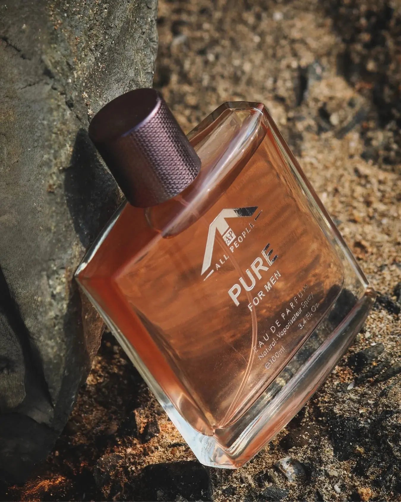 “All People ‘Pure’ perfume for men, set on sunlit sand beside a textured rock, showing a warm amber-toned glass bottle in a rugged outdoor setting.”