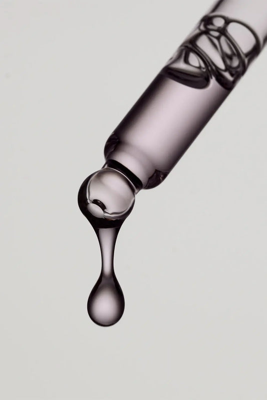 “Close-up of a serum dropper releasing a single drop of liquid, showing clear glossy texture against a plain light background.”