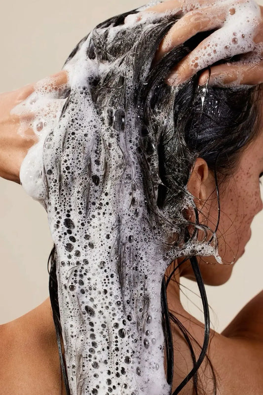 “Person massaging shampoo into wet hair, creating thick foam and lather while cleansing the scalp against a neutral background.”